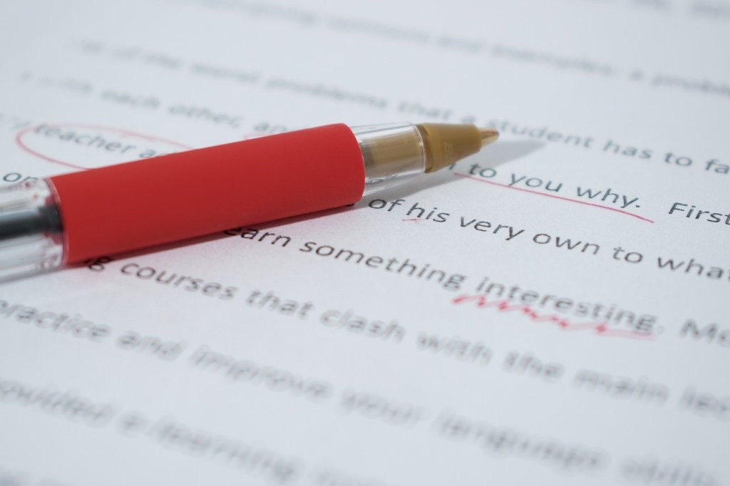 A picture of a red pen on a page of text with lots of red marks, representing the idea that the inability to write well is judged harshly