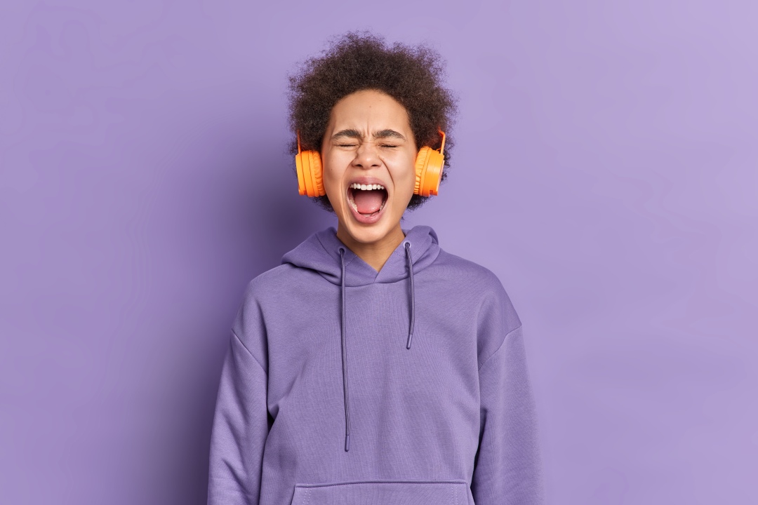 A woman wearing a purple hoodie standing against a purple background. The woman is wearing headphones, has her eyes shut, and is screaming. Introduces the topic of how angry I get when hearing poorly-written English lyrics in K-pop